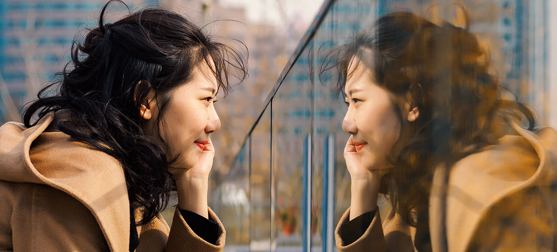 Frau sieht sich freundlich im Spiegelbild an. Durch eine wohlwollende Haltung uns selbst gegenüber können wir unser Selbstbewusstsein stärken