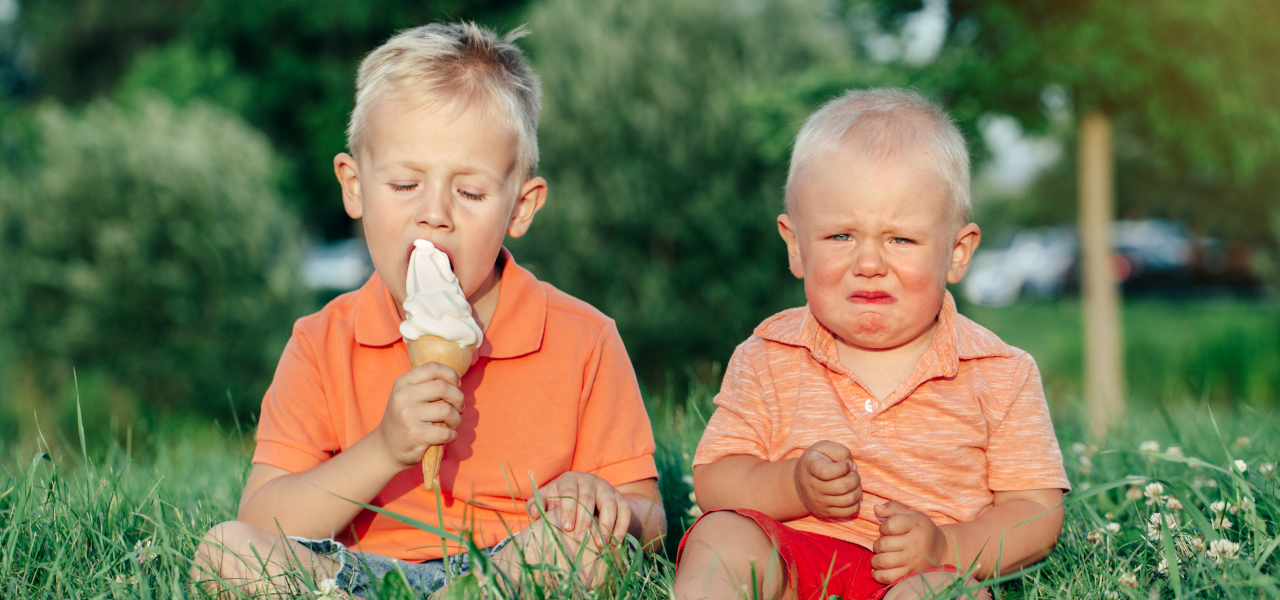 Zwei Kinder, der Junge links hat ein Eis in der Hand, das Kind rechts neben ihm hat kein Eis abbekommen. Es sieht unglücklich aus, man sieht ihm den Neid an