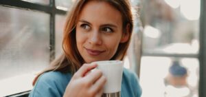 Eine junge Frau hält lächelnd eine Kaffeetasse in der Hand und schaut aus dem Fenster.