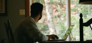 Ein Mann sitzt vor einem Laptop und schaut aus dem Fenster.