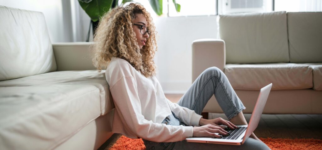 Eine Frau sitzt auf dem Boden an ein Sofa gelehnt und arbeitet an einem Laptop auf ihrem Schoß.